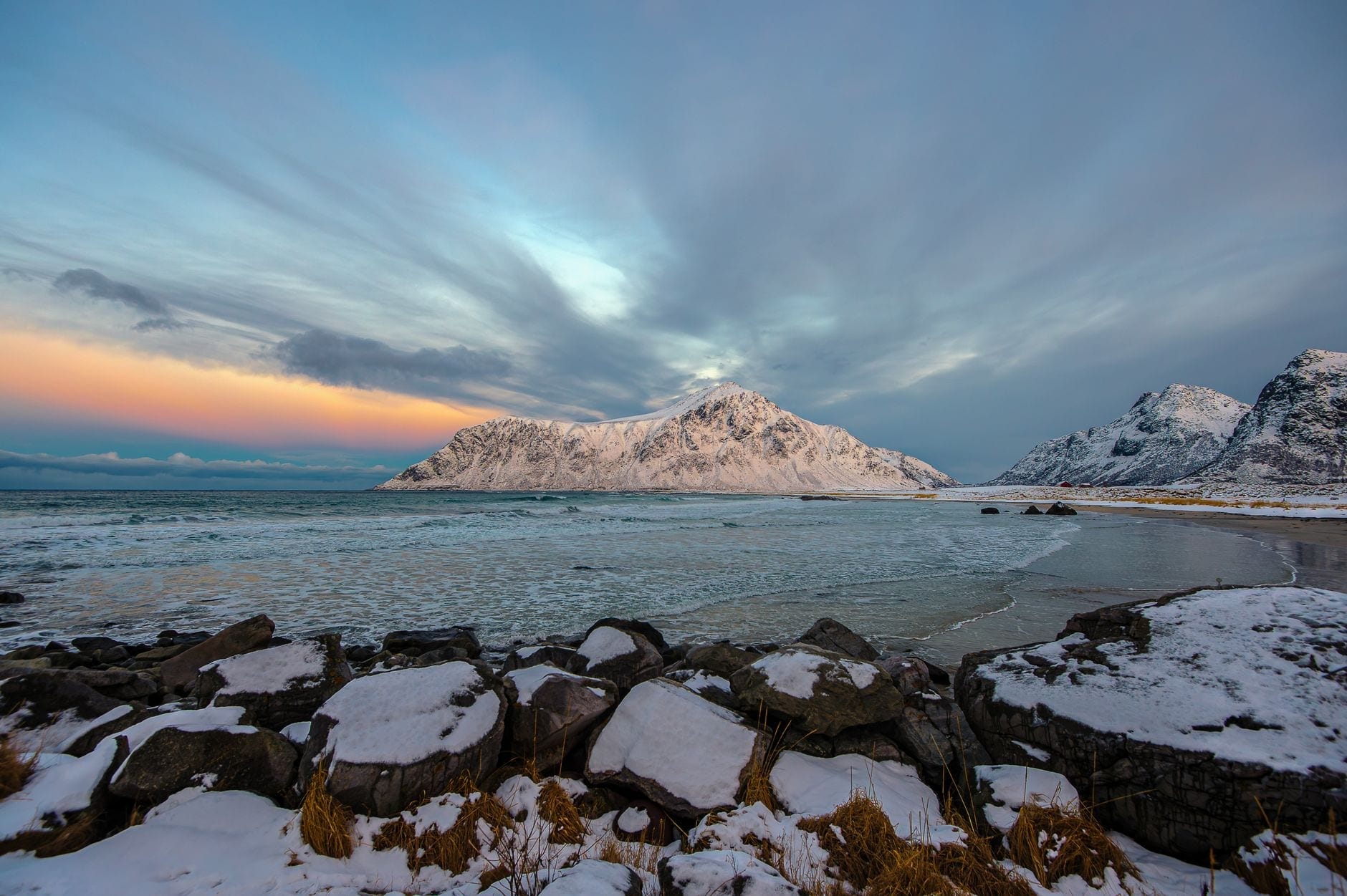 Snødekt strandlinje i Lofoten under en bratt arktisk fjelltopp og en stripet skumringshimmel