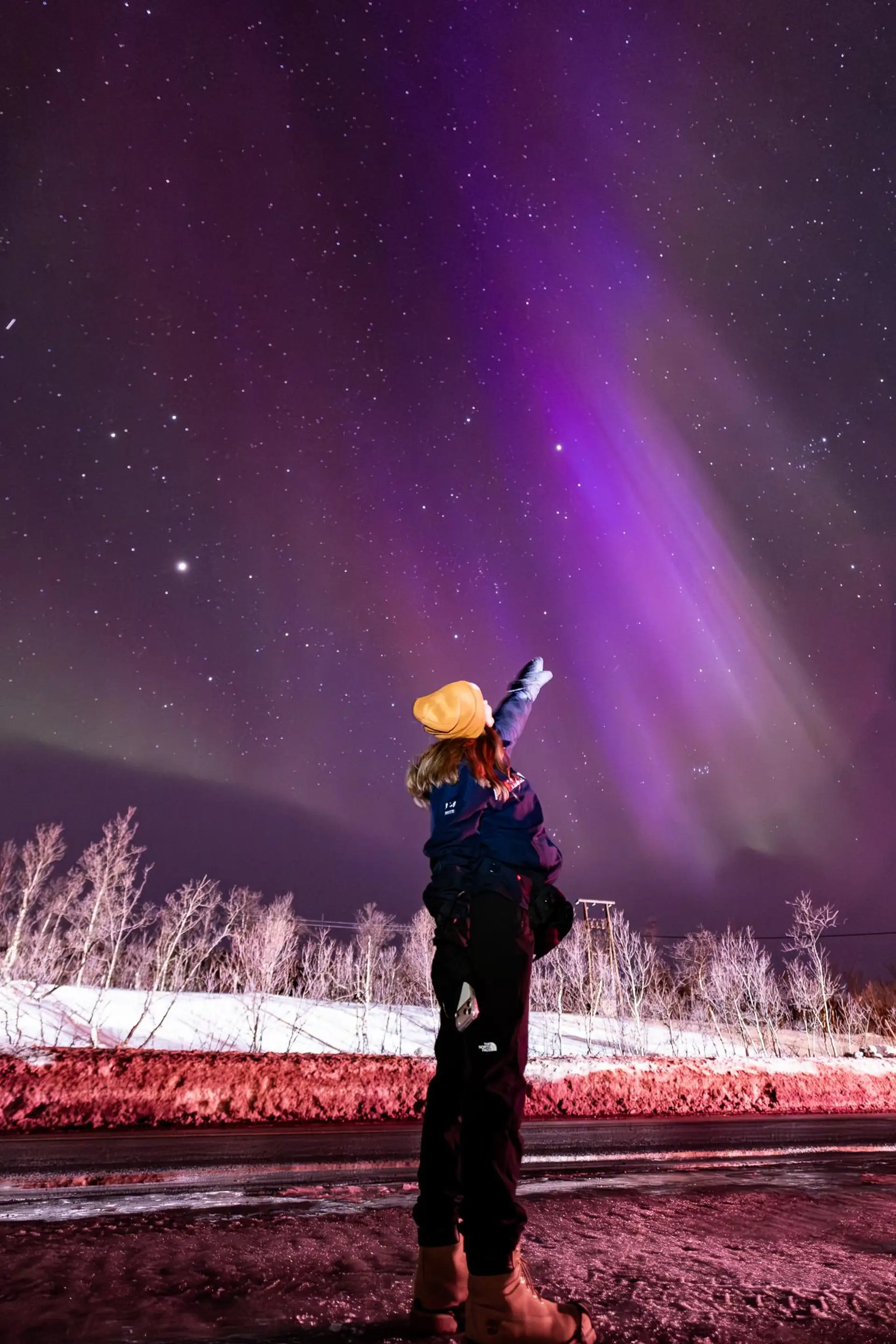 En gjest i gul lue peker opp mot fiolett nordlys over snødekt mark