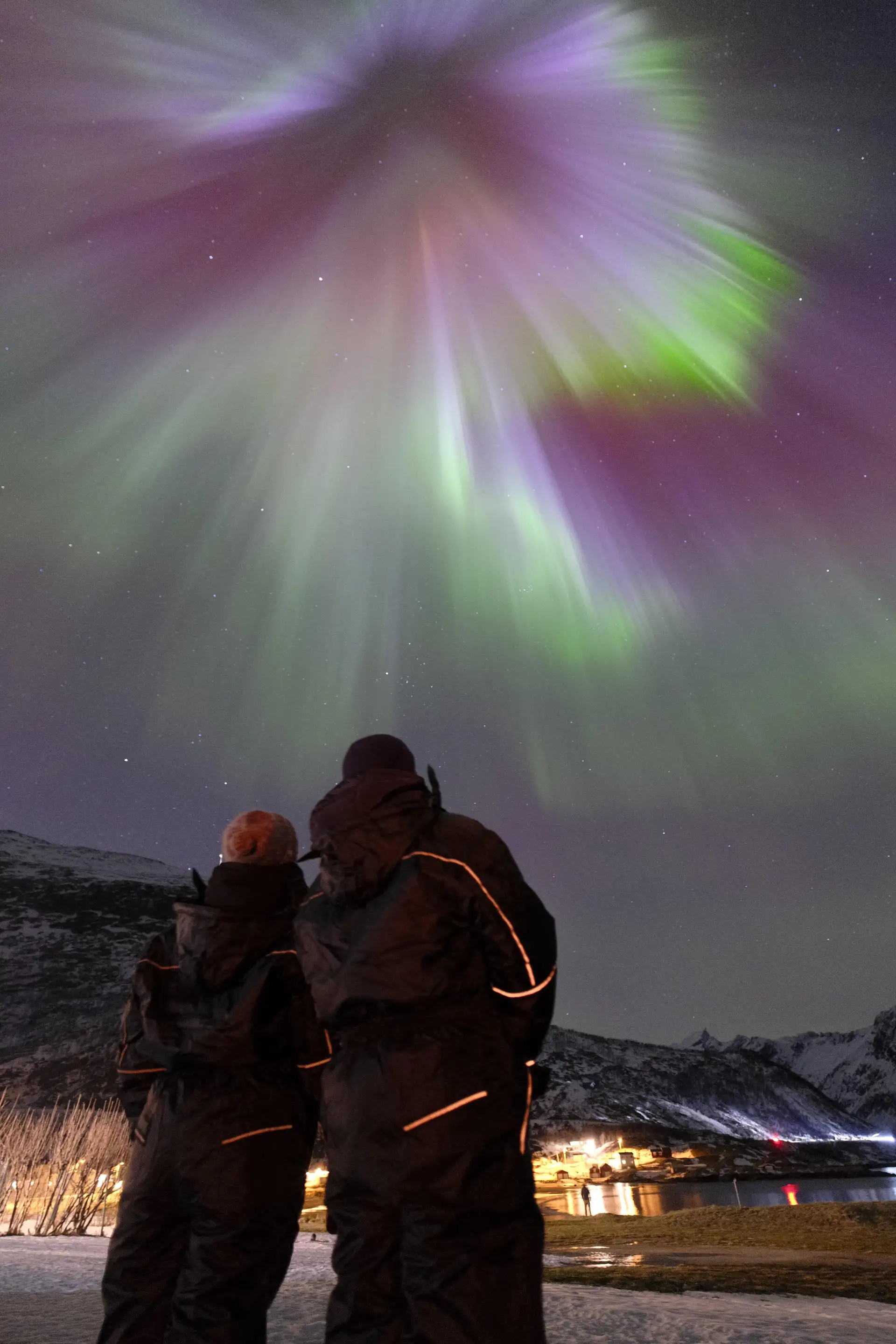 To gjester i termodresser ser på en grønn og lilla nordlyskorona over fjorden