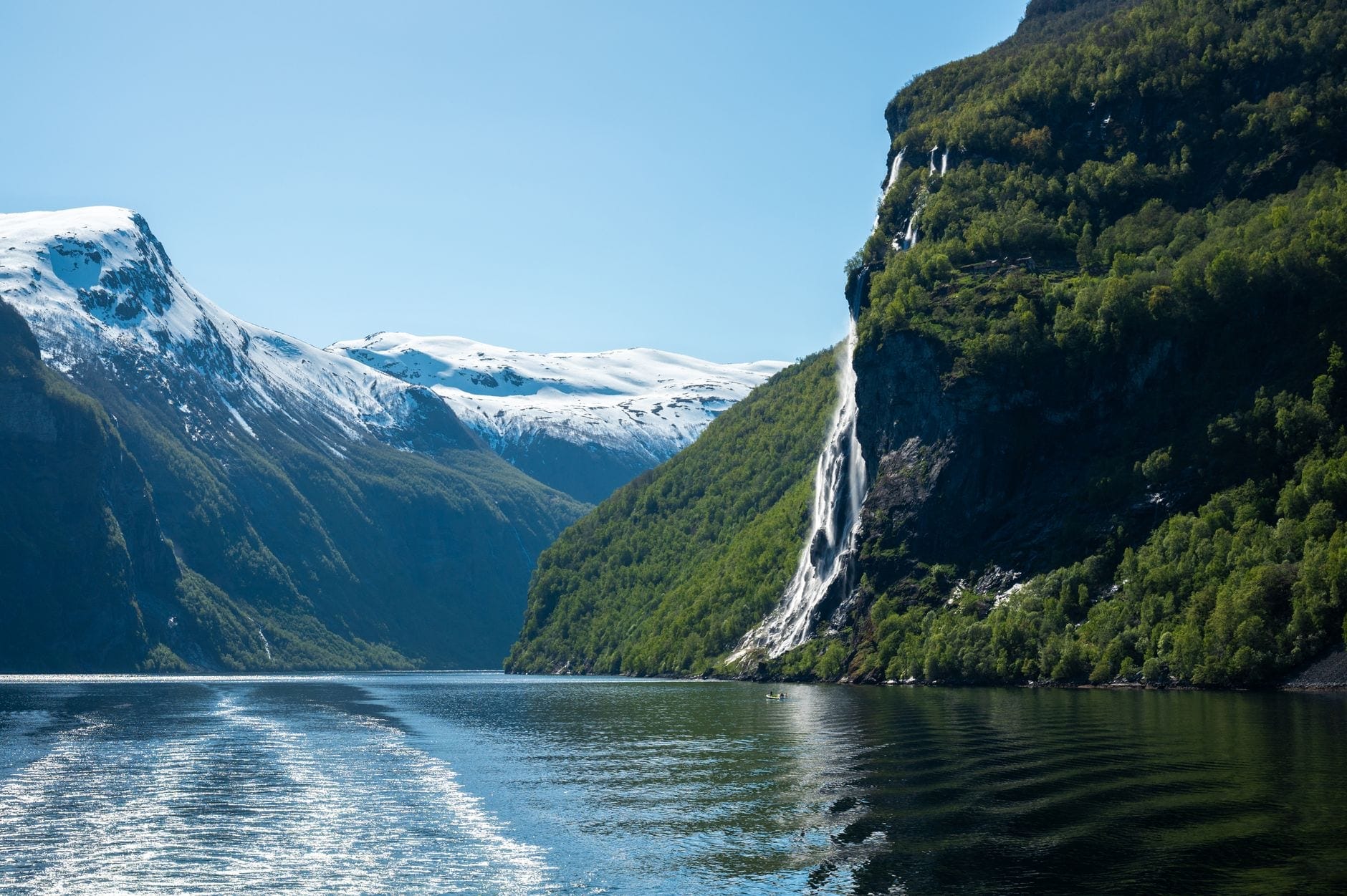 Snødekte fjordvegger og et vannfall sett fra kjølvannet til en båt