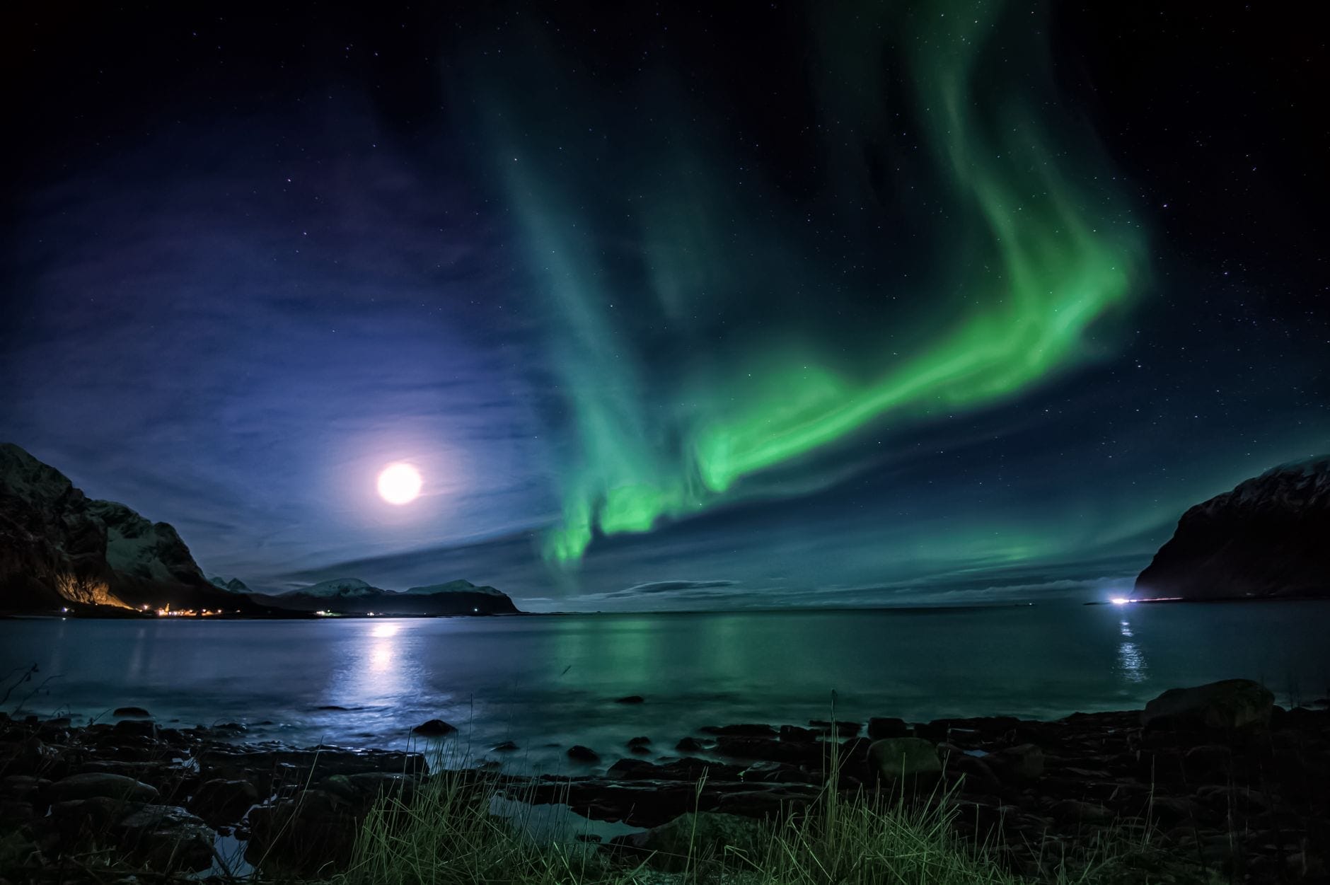 Nordlys og fullmåne over stille arktisk vann, med en steinete strandlinje i forgrunnen